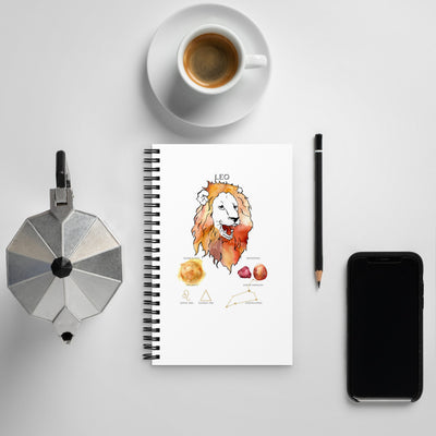 Notebook with a Leo zodiac illustration on a white surface with a cup, phone, and coffee press.