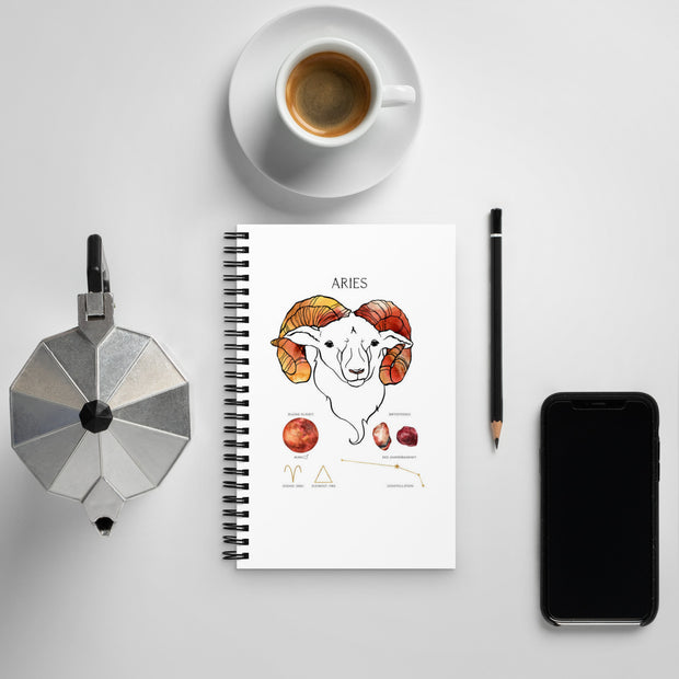 Notebook with Aries illustration, coffee cup, phone, and other items on a light gray surface