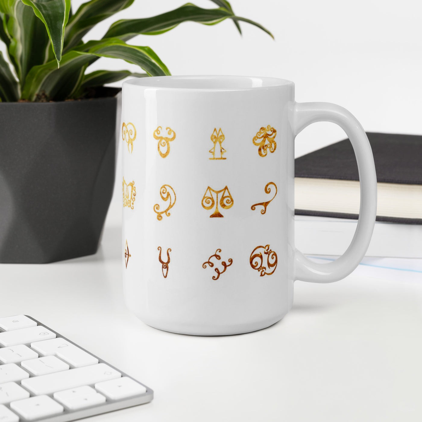 White mug with gold astrological symbol designs on a desk with a plant and books in the background