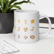 White mug with gold astrological symbol designs on a desk with a keyboard and plant.