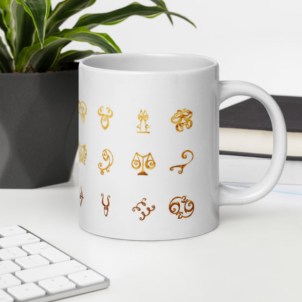 White mug with gold astrological symbol designs on a desk with a keyboard and plant.